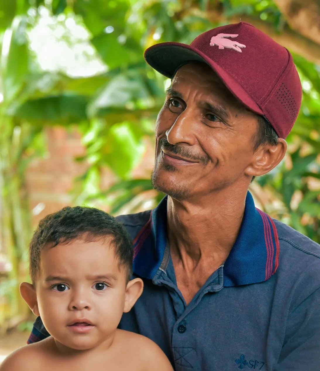Man in a burgundy baseball cap smiling to the left and holding his child.