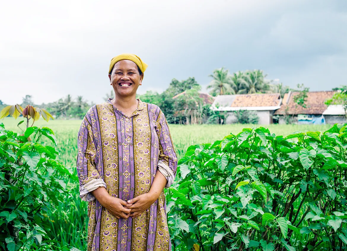 Aisyah, a woman in Indonesia smiling as she stands in a field of crops she grew.