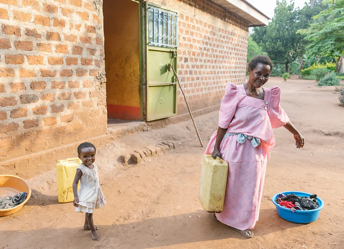 Woman holding a water vessel for laundry while standing next to a young girl outside of a brick building.